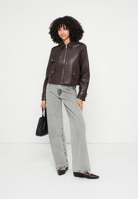 Hnedá krátka bunda z umelej kože s dvoma prednými vreckami, skombinovaná so svetlosivými džínsami s širokými nohavicami a čiernymi balerínami s špicatou špičkou. V ruke čierna kabelka.