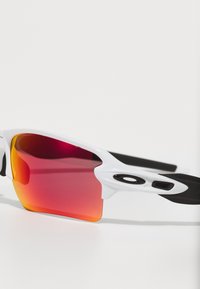 Białe sportowe okulary przeciwsłoneczne z zaokrąglonym kształtem, soczewkami w odcieniu czerwono-pomarańczowym oraz czarnymi gumowymi akcentami na ramionach dla lepszego uchwytu i komfortu.