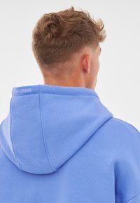 Felpa blu chiaro con cappuccio regolabile e dettagli a costine. Tessuto liscio con il marchio "Sandy Urban" sul retro del collo.