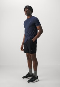 T-shirt de sport à manches courtes bleu marine, shorts noirs avec logo, chaussettes grises et chaussures de course noires. Le modèle se tient face à l'avant sur un fond clair.