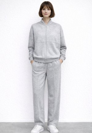 Kvinde står op ad en hvid væg iført en grå sweatshirt med lynlås, matchende bukser med hænderne i lommerne og hvide sneakers.