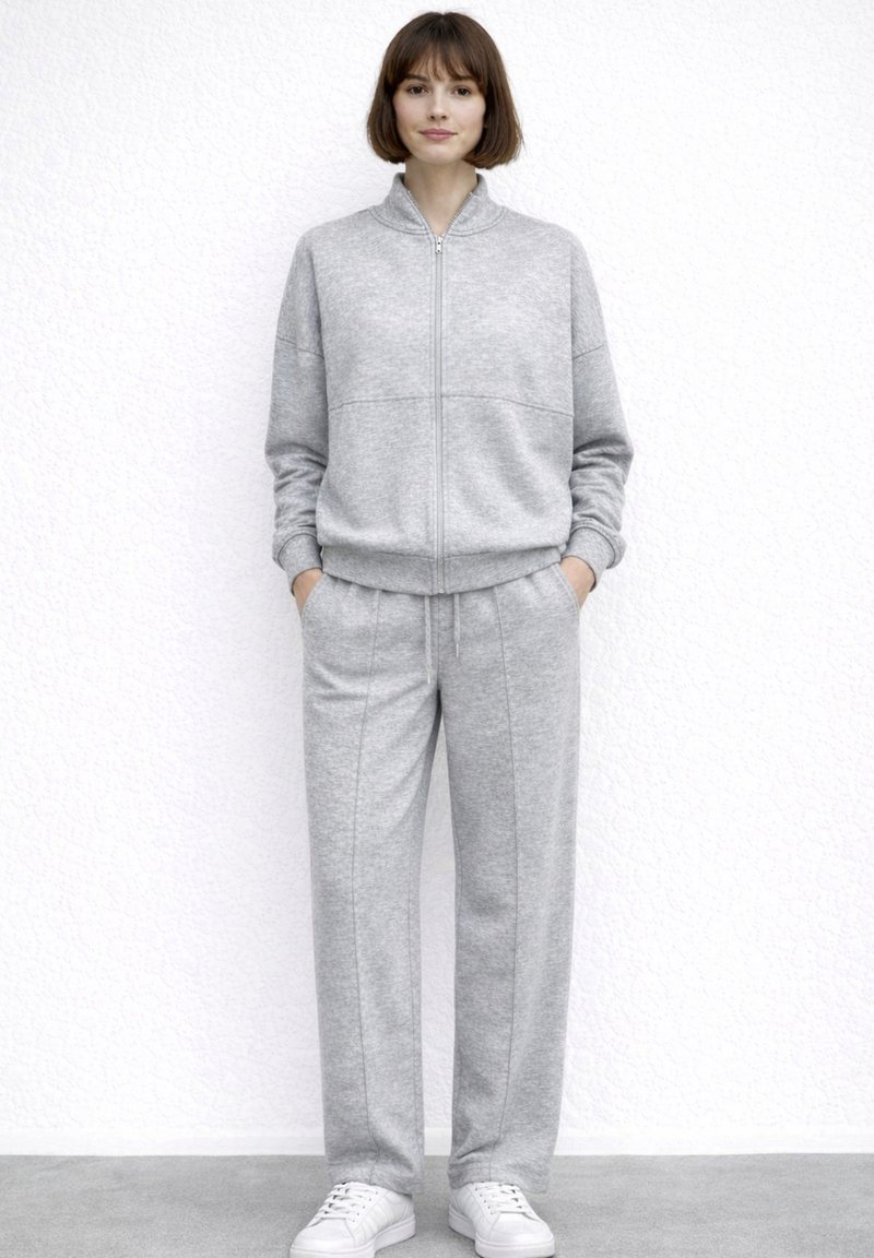 Kvinde står op ad en hvid væg iført en grå sweatshirt med lynlås, matchende bukser med hænderne i lommerne og hvide sneakers.