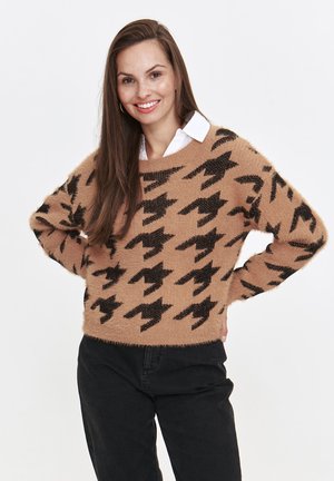 Beżowy sweter z czarnym wzorem w ekoskóry, miękka faktura i luźny krój. Noszony na białej koszuli z kołnierzykiem, z szerokim dekoltem.