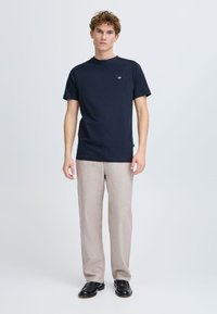Marineblauwe katoenen T-shirt met korte mouwen en een ronde hals, gecombineerd met beige broeken en zwarte schoenen. Heeft een klein logo op de borst.