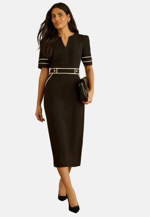 Schwarzes Midikleid mit kurzen Ärmeln, weißen Akzentlinien und einem taillierten Gürtel. Das Model hält eine schwarze Handtasche und trägt schwarze High Heels.