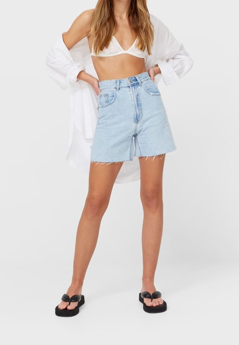 Femme portant un short en denim taille haute bleu clair, un bralette blanc, une chemise blanche oversize et des tongs noires à plateforme sur un fond blanc.