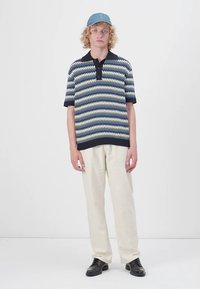Polo en maille présentant un motif en zigzag bleu, crème et marine, avec un col et des manches courtes, associé à un pantalon crème et des chaussures noires.