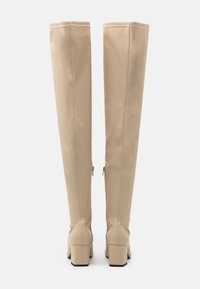 Botas beige por encima de la rodilla hechas de material liso con tacones cuadrados, que presentan un diseño sin costuras y detalles de cremallera en los lados.