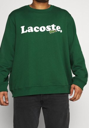 Grön bomullströja med rund halsringning, försedd med en vit "Lacoste."-logotyp och en broderad krokodil-logotyp på vänster bröst.