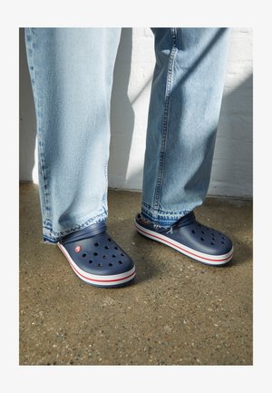 Marineblaue Clogs mit Belüftungslöchern, ausgestattet mit einer weiß-rot gestreiften Sohle, getragen mit hellblauen, gealterten Denim-Jeans.