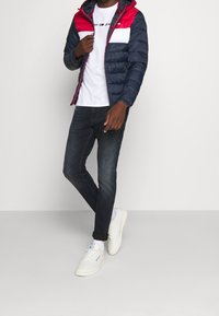 Herre pufferjakke med en marineblå krop, røde skuldre og en hvid brystpanel. Sammen med mørke jeans og hvide sneakers.