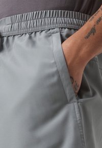 Main avec tatouages reposant dans la poche latérale d'un pantalon à taille élastique gris clair, vue en gros plan.