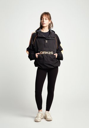 Black waterproof jacket with orange accents, front pocket, and logo "OAK25." Worn with black leggings and beige sneakers.