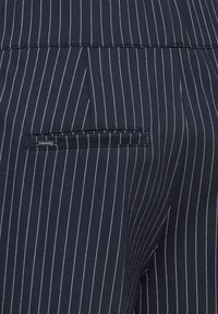 Tissu bleu marine à rayures fines mettant en avant une poche arrière avec un subtil détail de logo en métal. Le matériau est lisse avec une finition ajustée.