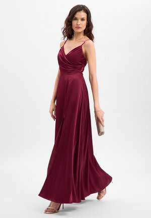 Maxi-jurk van bordeauxrood satijn met wikkel-effect, dunne bandjes, een vloeiende silhouet en een zijsplit; geaccentueerd met een gouden clutch.