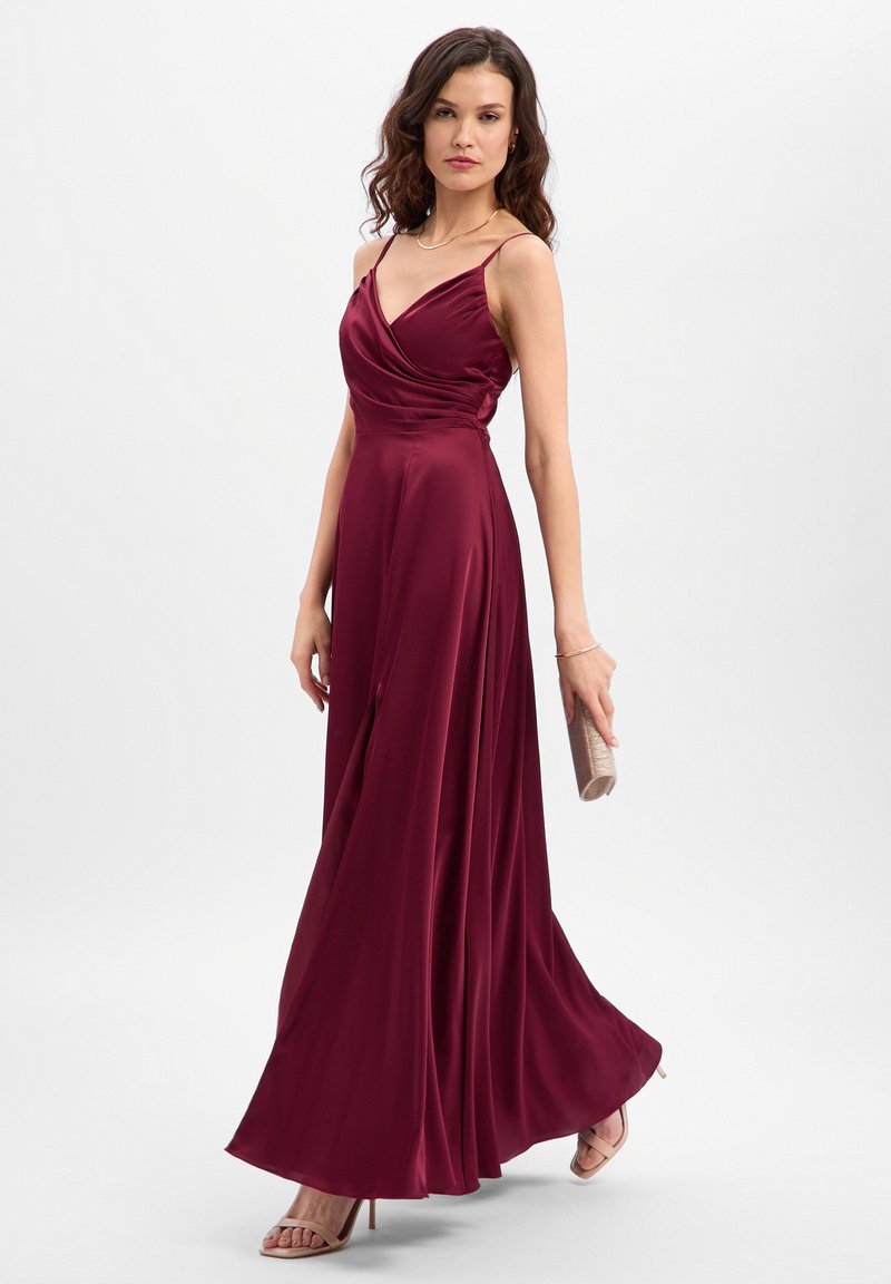 Maxi-jurk van bordeauxrood satijn met wikkel-effect, dunne bandjes, een vloeiende silhouet en een zijsplit; geaccentueerd met een gouden clutch.