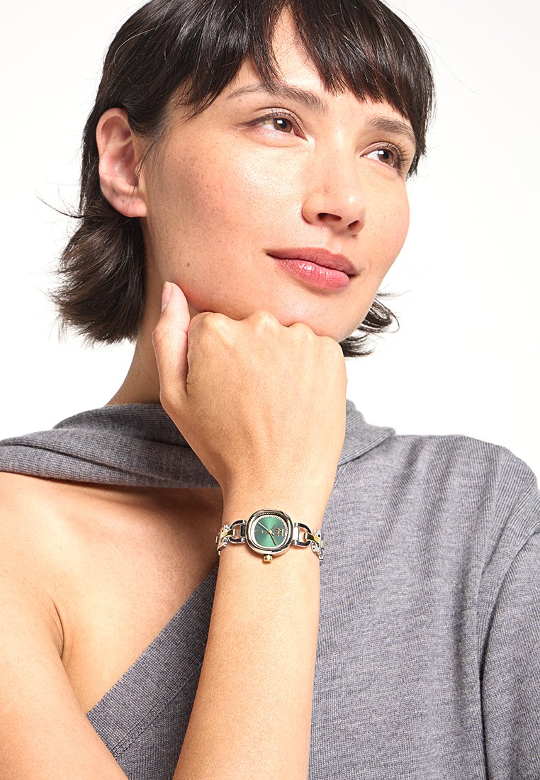 Orologio verde rotondo con finiture in argento e un bracciale metallico con maglie circolari. Il quadrante mostra dettagli e lancette dorate su uno sfondo verde.