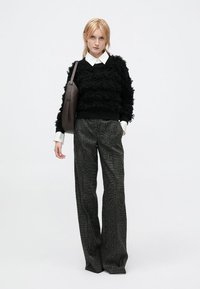 Schwarzer, strukturierter Pullover mit einem gerippten Saum, getragen über einem weißen Hemd mit Kragen. Dunkel gemusterte, weite Hose. Das Model trägt eine braune Umhängetasche.