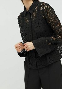 Vrouw in een zwarte bloemenkant blouse met lange mouwen en knopen, met brede manchetten, en zwarte broek tegen een effen achtergrond.