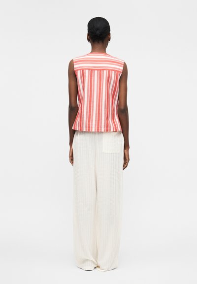 Γυναίκα με πίσω δεμένα μαλλιά, που φοράει κόκκινη και λευκή ριγέ αμάνικη μπλούζα και χαλαρό λευκό υφασμάτινο παντελόνι, όρθια, με την πλάτη προς τα έξω.