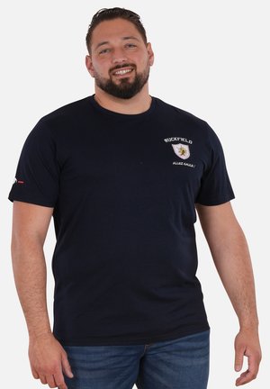 Homme souriant aux cheveux foncés et à la barbe, portant un t-shirt bleu marine Ruckfield et un jean bleu, debout devant un fond clair uni.