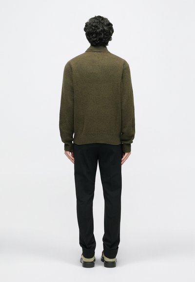 Olivgrüner Strickpullover mit hohem Kragen, kombiniert mit schwarzen Hosen und grünlichen Schuhen. Die Struktur ist gerippt und betont die Wärme.