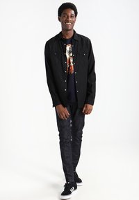 Camicia nera con bottoni e due tasche frontali, indossata sopra una t-shirt con grafica. Jeans scuri e sneaker nere. Modello in piedi su uno sfondo neutro.