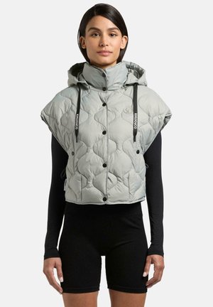 Frau trägt eine graue gesteppte ärmellose Kapuzenweste über einem schwarzen langärmeligen Oberteil und schwarzen Shorts, stehend vor einem schlichten Hintergrund.