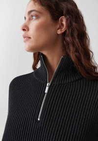 Junge Frau mit lockigem Haar, die einen schwarzen gerippten Pullover mit einem teilweise geöffneten silbernen Kragen trägt und nach links schaut.