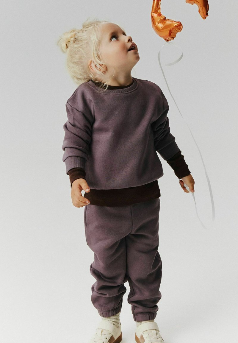Jeune enfant aux cheveux blonds en survêtement violet regardant un ballon orange en forme du chiffre trois avec une ficelle blanche.