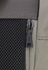 Siva tkaninska torba z črnim mrežnim panelom, ki vsebuje črn zadrgo s pravokotnim trakom in opozorilnim simbolom. Vidna teksturirana površina in šivi.