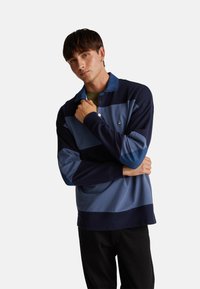 Polo triko s dlouhým rukávem v námořnické a modré barvě, zdobeno vodorovnými pruhy a kontrastním límečkem. Tkanina se zdá být hladká s mírnou strukturou.