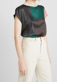 Mujer que lleva una blusa de manga corta con un patrón de bloques de color metida en pantalones de tiro alto color blanco roto con bolsillos frontales.