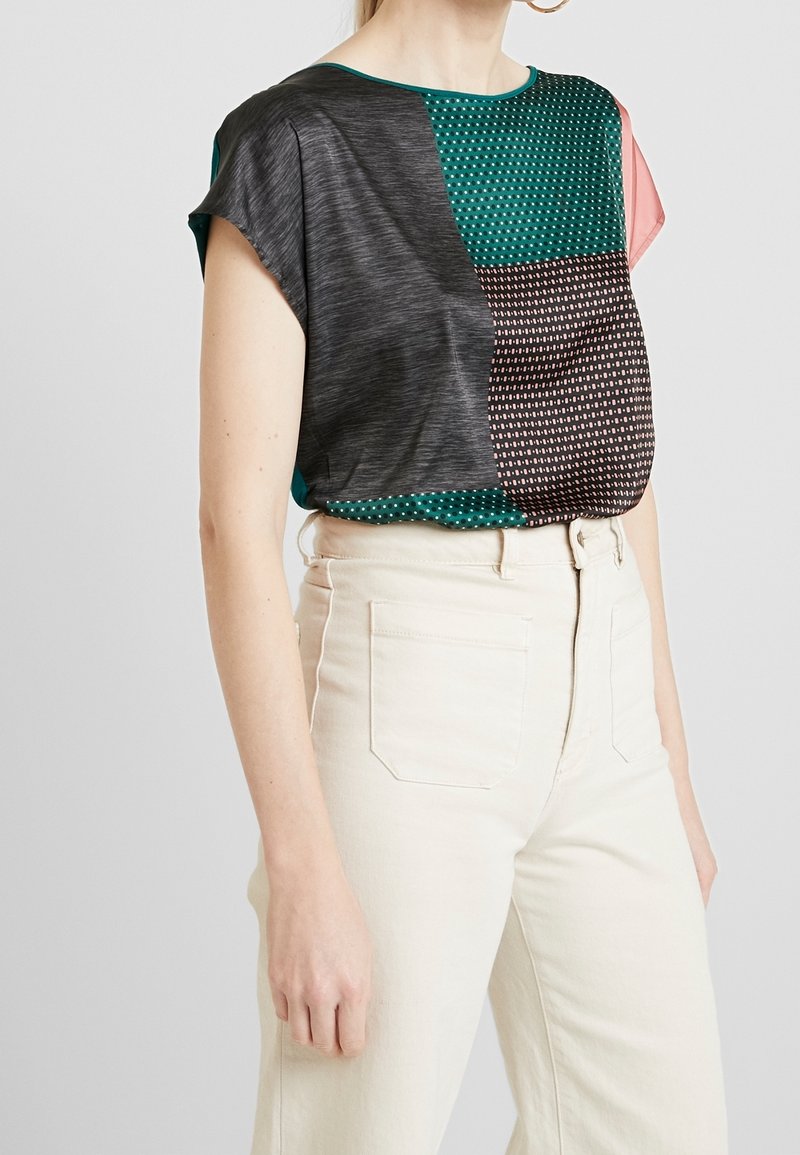 Mujer que lleva una blusa de manga corta con un patrón de bloques de color metida en pantalones de tiro alto color blanco roto con bolsillos frontales.