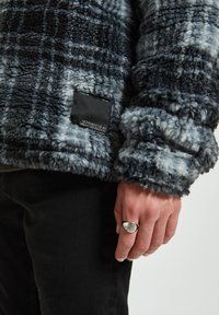 Ruka se stříbrným prstenem zobrazujícím symbol oka, oblečená v černé a šedé huňaté kostkované bundě a černých kalhotách.