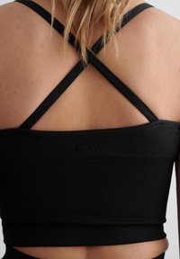 Musta ribbi krossitoppi, jossa on ristikkäiset hihnat selässä. Kangas on sileä ja tiukka, ja helmassa on huomaamaton logo.