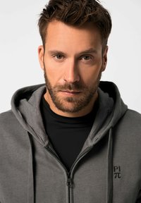 Sudadera gris con cremallera, hecha de una tela suave con capucha, que presenta acentos negros en la parte frontal y una ligera textura brillante.