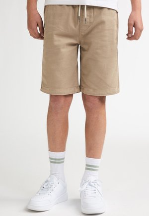 Pantalones cortos de algodón beige con cintura ajustable con cordón, bolsillos laterales y diseño de corte recto. Combinados con zapatillas blancas y calcetines a rayas.