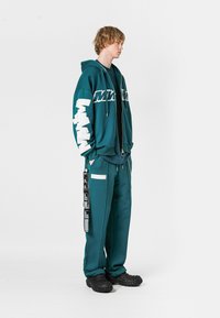 Sudadera teal con gráfico blanco y pantalones de chándal a juego. Las características incluyen cremalleras, bolsillos y puños elásticos, combinados con zuecos negros.