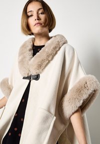 Vrouw in een crème cape met beige nepbont aan de kraag en mouwen, met een zwarte gesp over een zwarte jurk met bruine stippen.