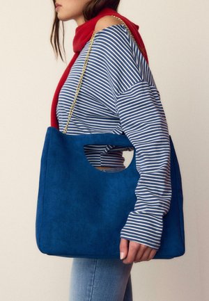 Frau in blau-weiß gestreiftem Oberteil mit rotem Schal, die eine große blaue Wildleder-Handtasche mit goldener Kettenriemen über der Schulter trägt.