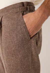 Pantalones marrones texturizados con un corte ajustado, que presentan un lazo para cinturón estrecho y una mano en el bolsillo, destacando el tejido.