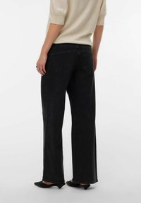 Personne portant un jean noir à jambes larges, un pull beige à manches courtes, des chaussures noires à talons bas, debout de dos face à la caméra.