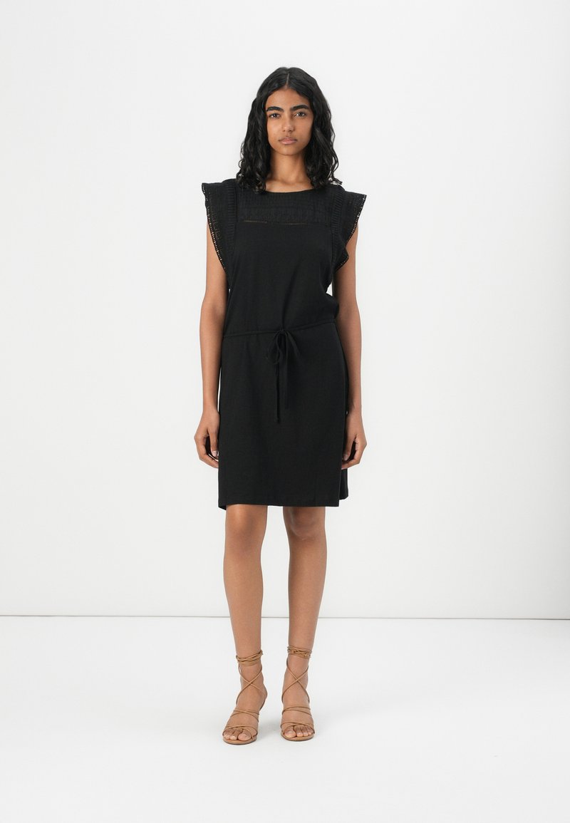 Black dress made of soft fabric, featuring ruffled sleeves and a drawstring waist. Flat sandals with crisscross straps complement the outfit.