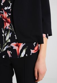 Blazer noir à manches trois-quarts, superposé à un chemisier à imprimé floral avec des lys roses et des feuilles vertes sur un fond sombre.