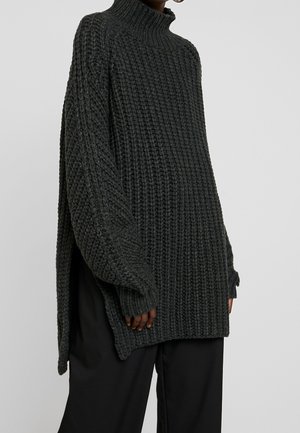 Oversize mørkegrå chunky striktrøje med høj hals og lange ærmer, båret med løstsiddende sorte bukser mod en ensfarvet baggrund.