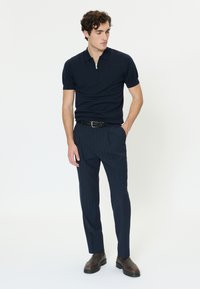 Uomo che indossa una polo blu navy a maniche corte con cerniera, pantaloni blu navy a righe sottili, cintura nera intrecciata e stivali marrone scuro, in piedi davanti a uno sfondo bianco.