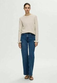 Maglione beige lavorato a maglia con polsini a balze, abbinato a jeans blu a gamba dritta. La modella indossa ballerine marroni, in piedi su uno sfondo chiaro.