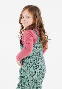 Tuta verde con macchie nere, indossato sopra un top in pile rosa. Presenta spalline regolabili, vita elastica e tasche laterali.