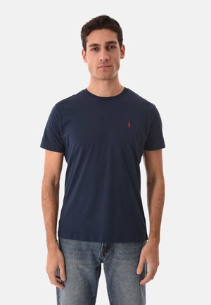 Uomo in piedi, volto rivolto in avanti, che indossa una maglietta blu navy semplice con un piccolo emblema a forma di peperoncino rosso sul petto e jeans blu.